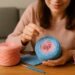 Eine Frau sitzt an einem Holztisch und beginnt, ihren Bobbel zu verarbeiten. Sie zieht den Faden vorsichtig aus der Mitte eines rosa-blauen Farbverlaufsbobbel, während daneben ein zweiter Bobbel und eine Häkelnadel liegen.